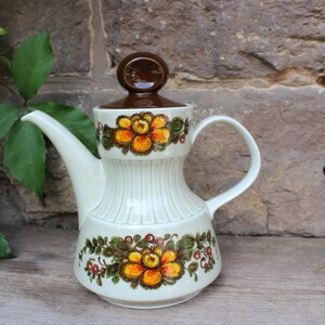 Könnte beinhalten: Eine weiße Teekanne aus Keramik mit braunem Deckel und Blumenmuster. Die Teekanne hat einen Ausguss und einen Henkel. Das Blumenmuster zeigt gelbe, orange und grüne Blumen.