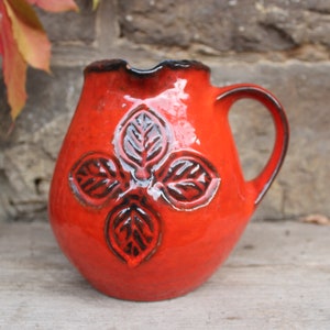 May include: A red ceramic pitcher with a black leaf design. The pitcher has a handle and a rounded body.