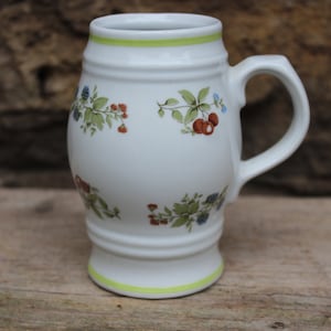 May include: White ceramic beer stein with a green rim and floral pattern featuring red berries, blue flowers, and green leaves.