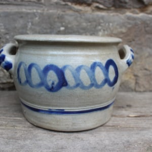 Könnte beinhalten: Ein Steinguttopf mit zwei Henkeln und einem Deckel. Der Topf ist hellgrau mit einem blauen Schlaufendesign in der Mitte und blauen Akzenten an den Henkeln. Der Topf steht auf einer Holzoberfläche.