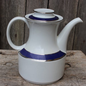Puede incluir: Tetera de cerámica blanca con una banda azul y plateada alrededor del centro. La tetera tiene una tapa con un borde plateado.