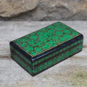 Puede incluir: Caja decorativa rectangular con base y tapa negras, adornada con un patrón floral verde y detalles dorados. La caja se encuentra sobre un fondo natural y texturizado.