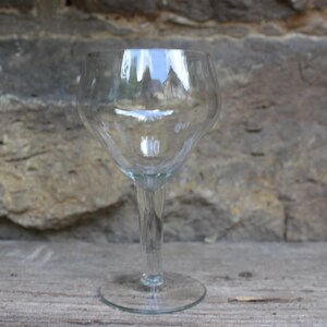 May include: A clear glass wine goblet with a wide bowl and a long stem. The goblet is sitting on a wooden surface.