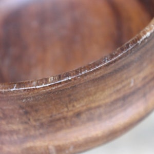 Puede incluir: Primer plano de un cuenco de madera con acabado marr&oacute;n oscuro. El borde del cuenco tiene un borde m&aacute;s claro y texturizado. La veta de la madera es visible, lo que se suma a la est&eacute;tica natural. La forma del cuenco es redonda y lisa.