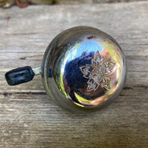 Könnte beinhalten: Eine runde Fahrradklingel aus poliertem Chrom mit einem eingravierten Kirschblütenmotiv auf der Oberseite. Die Klingel verfügt über einen seitlich angebrachten schwarzen Kunststoffhebel, der für die manuelle Bedienung einen hellen Klang erzeugt.