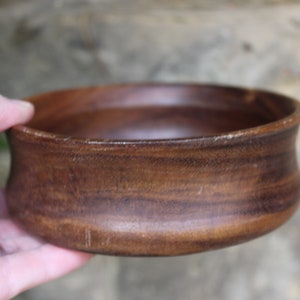 Puede incluir: Un cuenco de madera hecho a mano con un rico acabado marr&oacute;n oscuro. El cuenco tiene un dise&ntilde;o ancho y poco profundo con un borde ligeramente ensanchado. La veta de la madera es visible, lo que a&ntilde;ade encanto r&uacute;stico. El cuenco es sostenido por una mano.