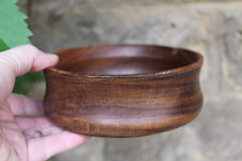 Puede incluir: Un cuenco de madera hecho a mano con un rico acabado marr&oacute;n oscuro. El cuenco tiene un dise&ntilde;o ancho y poco profundo con un borde ligeramente ensanchado. La veta de la madera es visible, lo que a&ntilde;ade encanto r&uacute;stico. El cuenco es sostenido por una mano.