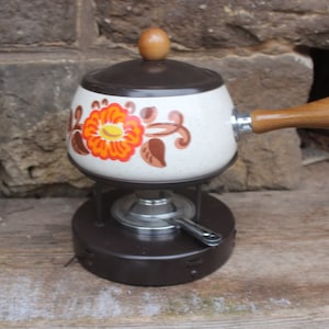 May include: A vintage fondue pot with a white base and brown lid, decorated with orange and brown floral designs. The pot has a wooden handle and sits on a metal stand with a burner.