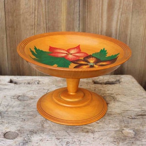 May include: A wooden pedestal bowl with a hand-painted floral design. The bowl is painted with red, orange, and green flowers on a light brown background.
