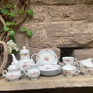 May include: A vintage tea set with a floral design. The set includes a teapot, sugar bowl, creamer, plates, and teacups. The white porcelain pieces are decorated with purple and yellow flowers.