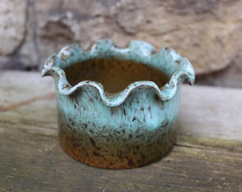 May include: A small, teal-colored ceramic planter with a scalloped rim. The planter has a brown glaze with a speckled pattern.