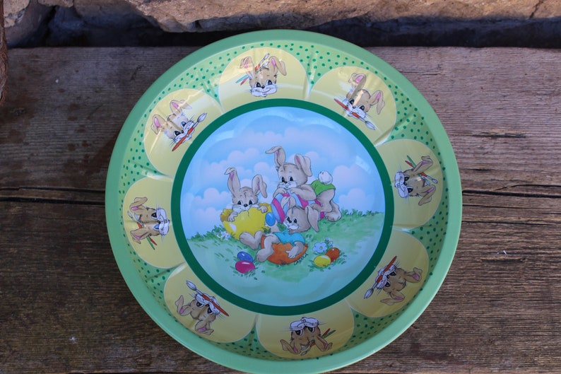 May include: A green metal bowl with a yellow rim and a floral design. The bowl features a cartoon image of three bunnies holding Easter eggs. The bunnies are surrounded by green grass and blue clouds.