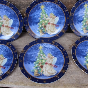 May include: Set of six decorative plates with a blue and gold rim. Each plate features a winter scene with an angel decorating a Christmas tree. The angel is wearing a white gown and has white wings.