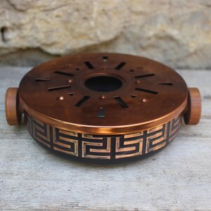 Op de afbeelding: Een ronde, koperkleurige wierookbrander met een geometrisch ontwerp rond de basis. De bovenkant heeft uitgesneden vormen en twee houten handvatten. De wierookbrander is een decoratief item.