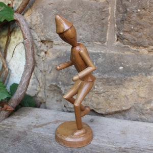 May include: A wooden figurine of a person wearing a pointed hat, with a whimsical, stylized design. The figurine is painted in a natural wood tone and is posed in a dynamic, energetic stance.