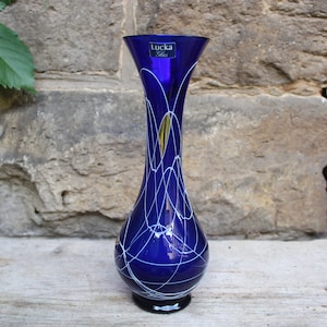 May include: A cobalt blue glass vase with white and gold abstract lines. The vase has a narrow neck and a rounded base. The vase is labeled "Lukka Glas".