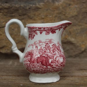 Puede incluir: Una jarra de crema de cerámica blanca con un diseño floral rojo y blanco. La jarra tiene un asa y un pico. El diseño presenta una escena de caza con jinetes a caballo.