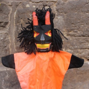 Könnte beinhalten: Eine Handpuppe mit einem schwarzen Holzgesicht, roten Hörnern und gelben Zähnen. Die Puppe trägt ein orangefarbenes Kostüm mit schwarzen Ärmeln.