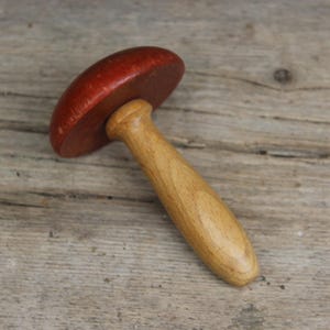 Może przedstawiać: Drewniany przedmiot w kształcie grzyba z czerwonym kapeluszem i jasnobrązowym trzonkiem. Grzyb leży na zwietrzałej drewnianej powierzchni, ukazując naturalne usłojenie i fakturę drewna.