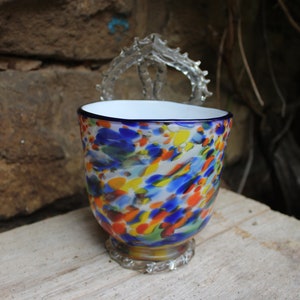 Könnte beinhalten: Eine bunte Glas-Tasse mit weißem Innenraum und blauem Rand. Die Tasse hat ein gesprenkeltes Muster in Blau, Gelb, Orange und Rot. Die Tasse steht auf einer Holzoberfläche.