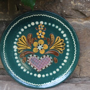Könnte beinhalten: Ein grüner Keramikteller mit weißem Rand und einem bemalten Blumendesign. Das Design zeigt ein Herz mit einem rosa-weiß karierten Muster, umgeben von gelben und braunen Blumen.