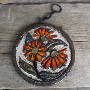 Könnte beinhalten: Ein runder Wandbehang aus Keramik mit einem floralen Design. Die Blumen sind orange, rot und gelb mit braunen und blauen Blättern. Der Wandbehang hat eine schwarze Metallkette.