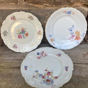 May include: Three antique white porcelain plates with floral designs. The plates feature scalloped edges and are decorated with colorful flowers in pink, blue, orange, and yellow. The plates are arranged on a weathered wooden surface.