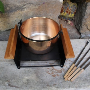 Può includere: Un pentolino per fondue in rame con manici in legno poggia su un supporto in metallo nero. Quattro lunghe forchette per fondue con manici in legno sono disposte su una superficie in legno.