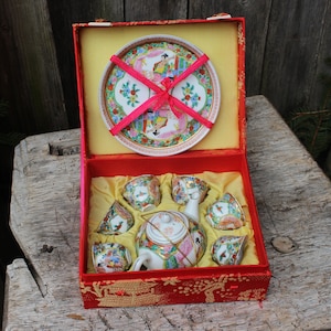 May include: A red and gold Chinese tea set in a red box with a floral design. The set includes a teapot, six cups, and a saucer. The box is lined with red satin.