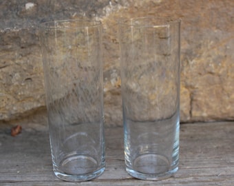 May include: Two clear glass drinking glasses with a textured swirl design. The glasses are empty and sitting on a wooden surface.