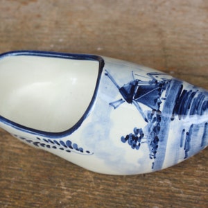 Könnte beinhalten: Ein weißer Keramik-Klomp mit blau-weißen Delftware-Designs. Der Klomp zeigt eine Windmühle und eine Landschaftsszene.