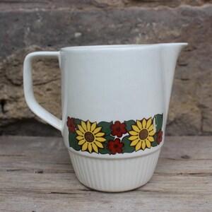 Könnte beinhalten: Weißer Keramik-Krug mit einem Blumendesign mit gelben Sonnenblumen und roten Blumen. Der Krug hat einen Henkel und einen leicht ausgestellten Rand.