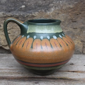 Könnte beinhalten: Keramikkrug mit Henkel, mit dunkelgrünem Rand und Henkel, einem hellgrünen Band und einem braunen Körper mit orangefarbenen und weißen dekorativen Elementen. Der Krug steht auf einer Holzoberfläche.