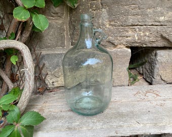 May include: A clear, light green glass jug with a rounded body, a narrow neck, and a small integrated handle near the top. The vessel features a smooth, transparent surface and a thick, sturdy base, showcasing a classic design for liquid storage.