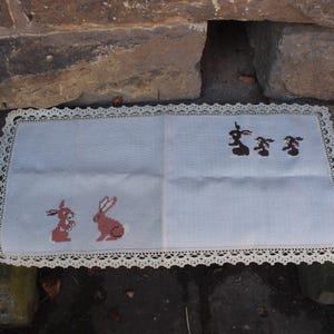 Op de afbeelding: Een wit linnen doily met een gehaakte kantzoom heeft geborduurde afbeeldingen van konijnen en hazen. De doily ligt op een houten oppervlak.