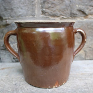 May include: A brown ceramic pot with two handles. The pot has a glossy finish and a slightly lighter rim. The background features a stone wall and a wooden surface.
