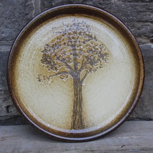 Könnte beinhalten: Ein runder Keramikteller mit braunem Rand und hellbraunem Glase. Ein stilisierter Baum ist in Braun auf den Teller gemalt.