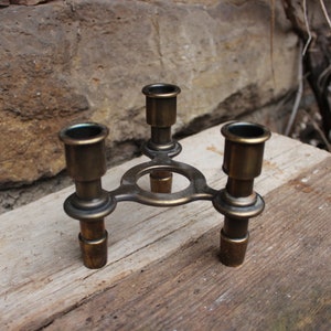 May include: A brass candle holder with three arms, each with a round cup for holding a candle. The holder has a circular base and is sitting on a wooden surface.