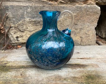 May include: A teal-coloured glass jug with a clear glass handle. The jug has a round body and a textured surface, with a dark blue interior. The jug is sitting on a wooden surface.
