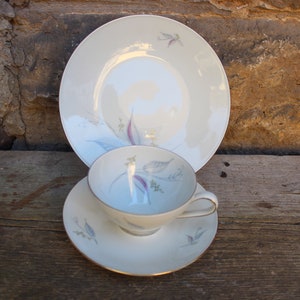 May include: A vintage porcelain teacup, saucer, and plate set. The set features a cream-colored base with a delicate floral design in shades of purple, blue, and green. The edges of the cup, saucer, and plate are trimmed with a gold band.