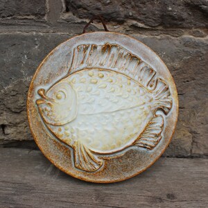 May include: A ceramic wall plaque featuring a white fish with brown details, set against a brown and white background.