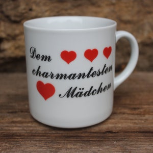 May include: White ceramic mug with a handle, featuring the German text "Dem charmantesten Mädchen" and four red heart symbols. The mug is set against a rustic wooden surface.