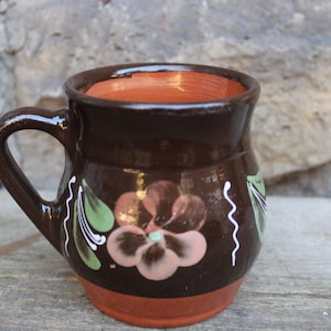 Könnte beinhalten: Eine braune Keramiktasse mit einem bemalten Blumendesign. Die Tasse hat einen abgerundeten Körper und einen Henkel. Das Blumendesign zeigt eine rosa Blume mit braunen und grünen Akzenten.