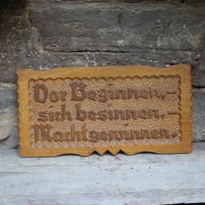 Könnte beinhalten: Rechteckiges Holzschild mit geschnitztem Text in dekorativer Schrift. Das Schild ist hellbraun mit einem strukturierten Rand und dem Text "Der Beginn, sich besinnen, Macht gewinnen."