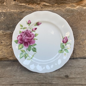 Könnte beinhalten: Weißer Keramikteller mit gewelltem Rand, verziert mit einer großen, leuchtend rosa Rose und kleineren Knospen. Grüne Blätter und zarte weiße Akzente ergänzen das florale Design. Ein klassischer, dekorativer Teller.