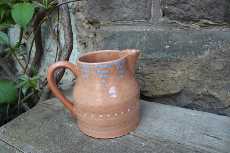 May include: A terracotta pitcher with a blue and white polka dot design. The pitcher has a wide base and a rounded body.
