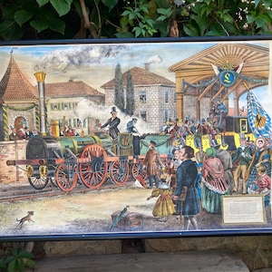 May include: A vintage-style educational wall chart featuring a colorful illustration of a 19th-century steam locomotive at a train station. The scene depicts passengers in period clothing, a large stone building, and a blue flag with a gold crest.