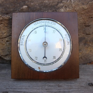 May include: A vintage barometer with a white face and a dark wood frame. The face has a needle pointing to the current pressure, with the words "Regen" and "Schon" on either side. The barometer is likely used to predict weather conditions.