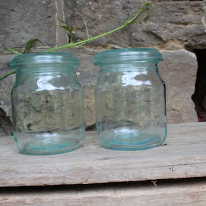 Op de afbeelding: Twee heldere glazen potten met een lichtblauwe tint en metalen deksels. De potten staan op een houten oppervlak.