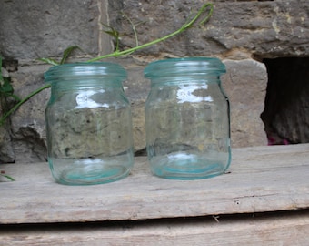 May include: Two clear glass jars with a light blue tint and metal lids. The jars are sitting on a wooden surface.
